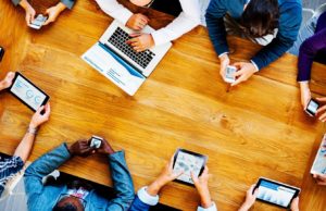 Co-workers sitting around a wooden table with electronic devices in hand - Search Influence