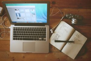 Apple laptop next to a notebook and a camera on a wooden surface