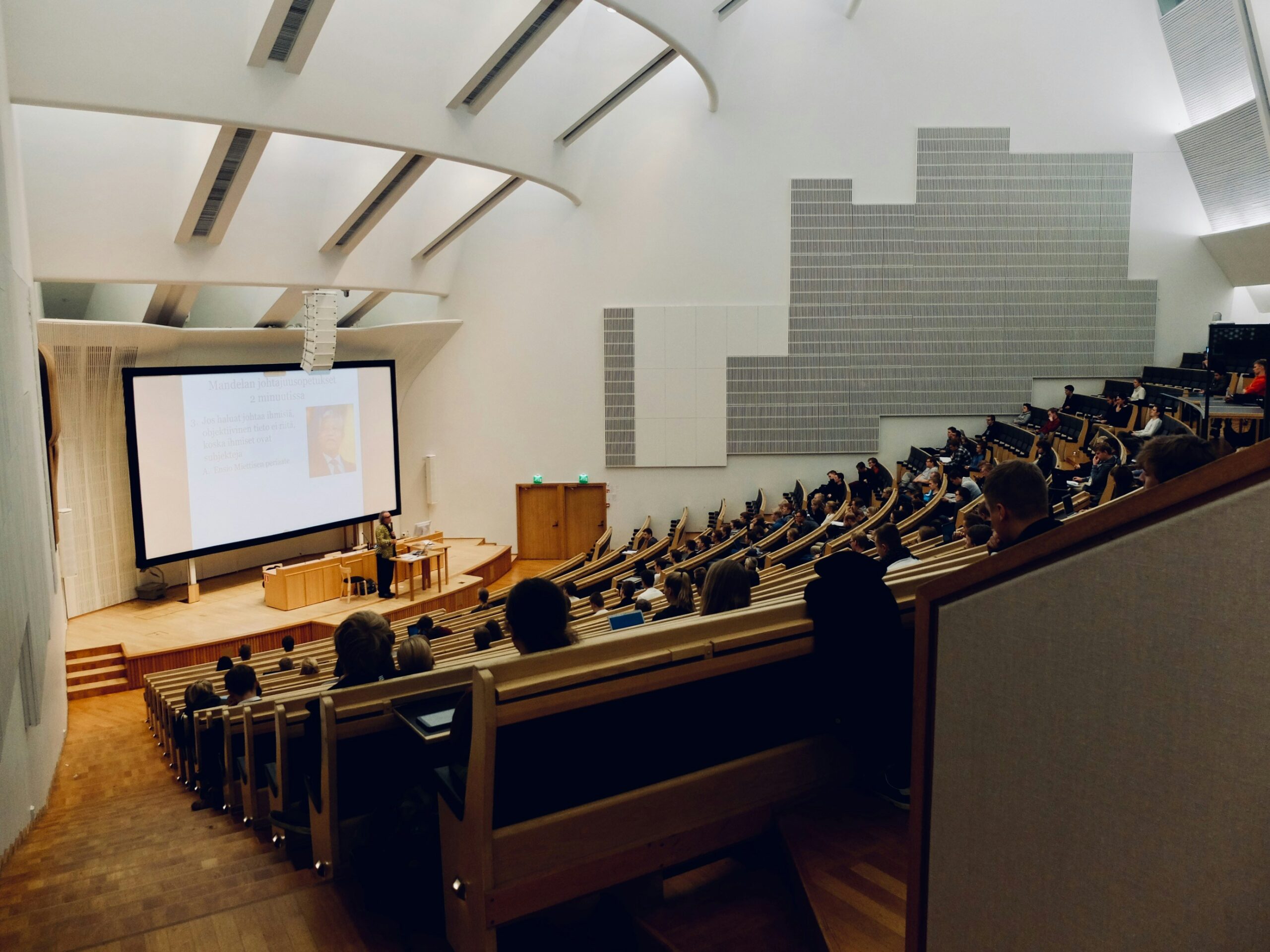 Students attending a lecture in a large university auditorium while a professor presents at the front.